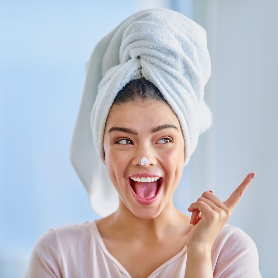Mujer sonriente con toalla en la cabeza aplicando crema facial en la nariz